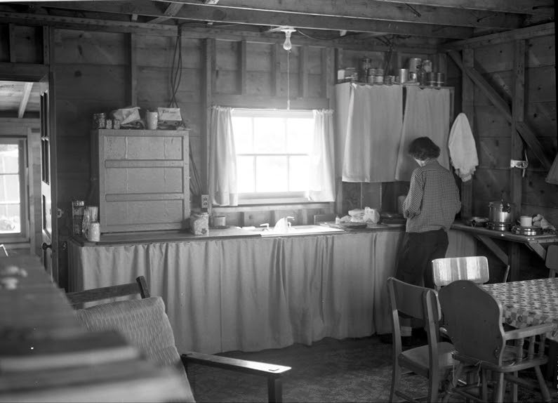 Then: Elsie at work in the kitchen around 1950, with curtains hung under the counter and the studs still bare — the interior wallboard did not go up until fourteen years after the cabin was built. Wes's History, photo 15.